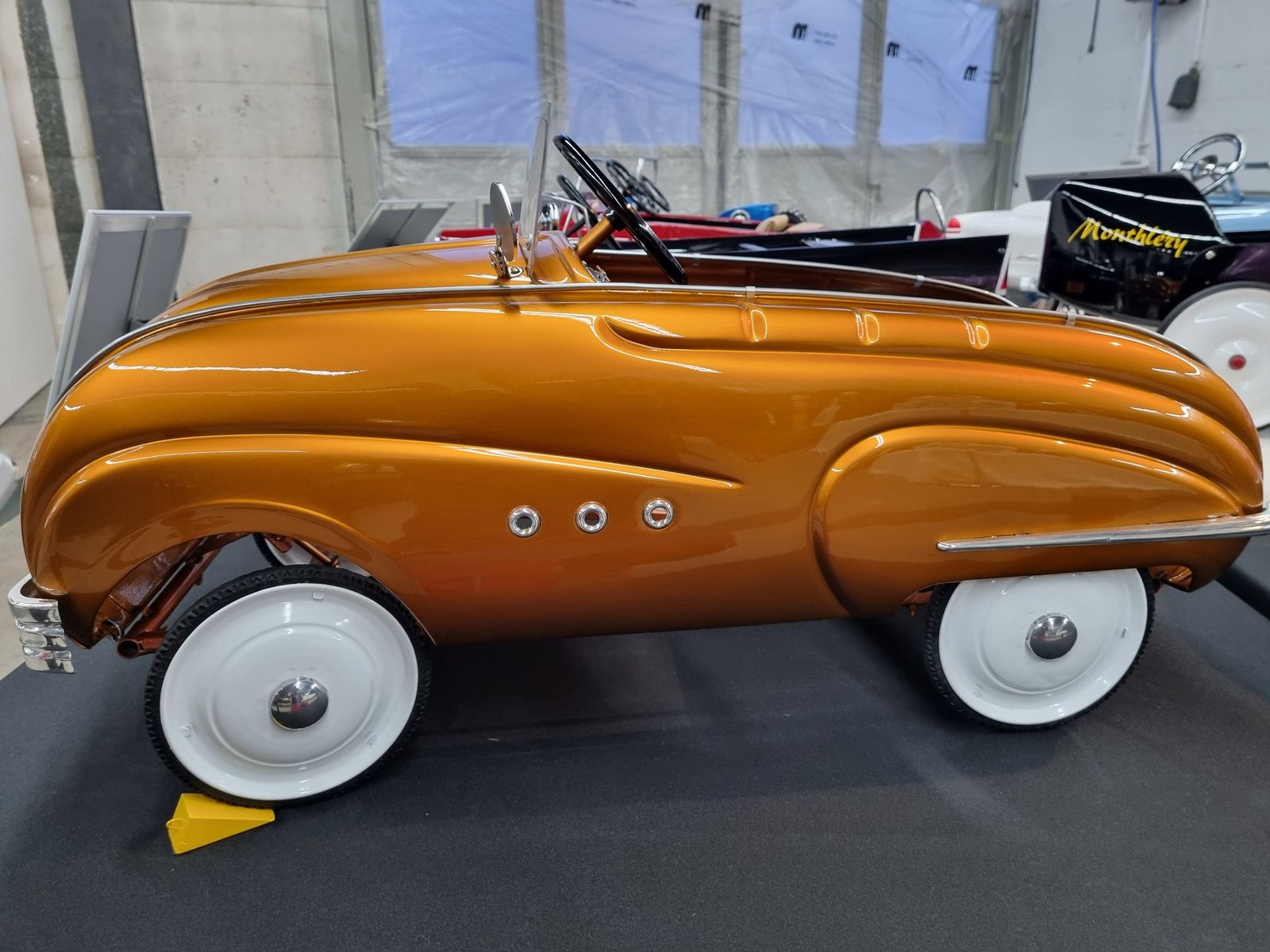 In einem Raum steht ein goldenes Tretauto mit weißen Rädern.